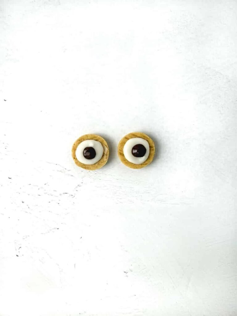 Two small vanilla sandwich cookie halves on a white surface, each topped with melted white and brown chocolate to create owl eyes for decorating