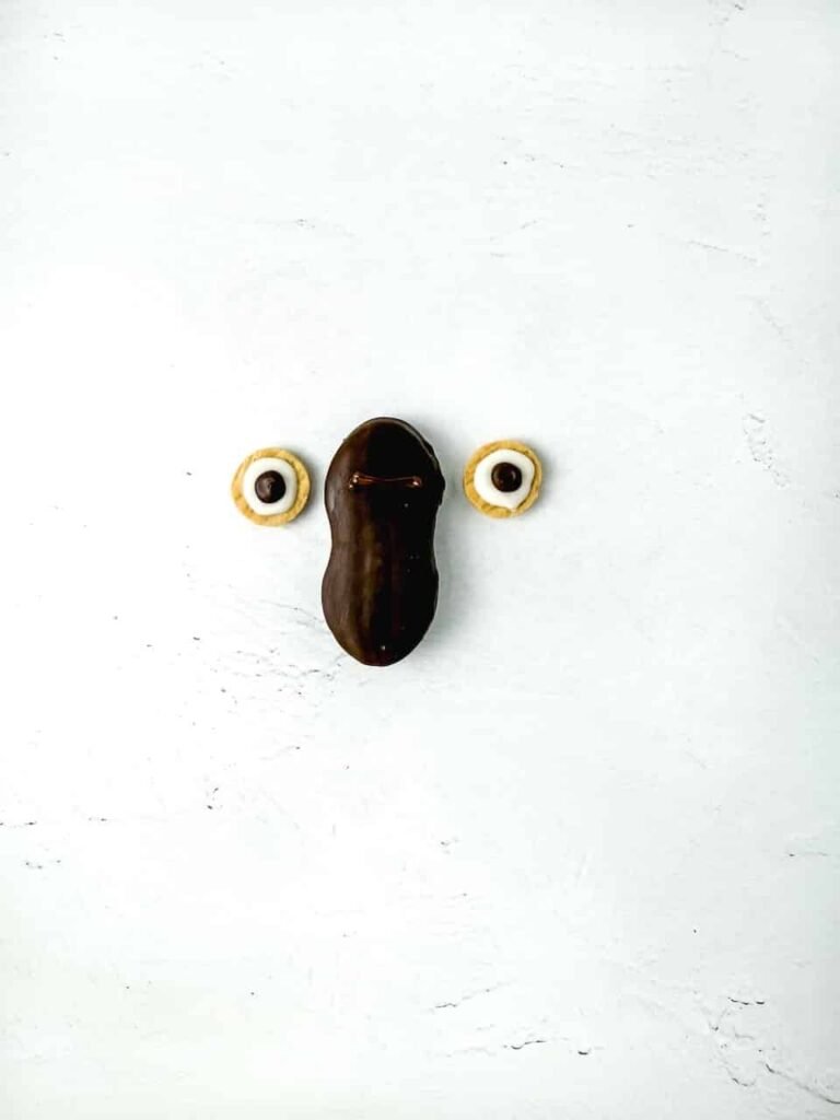 Chocolate-coated cookie on a white surface with two decorated mini vanilla cookie halves beside it, showing how to attach the owl eyes for the Nutter Butter owl cookies.