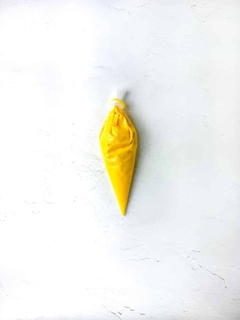A piping bag filled with melted yellow chocolate resting on a white surface, prepared for piping small beaks onto the cookies during the decorating step.