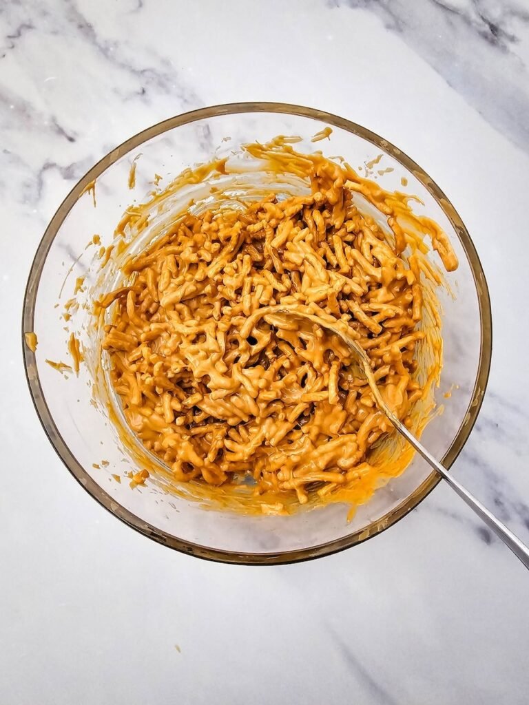 Chow mein noodles coated with melted butterscotch and peanut butter being stirred together in a glass mixing bowl.