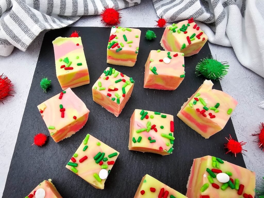 Colorful swirled Christmas fudge squares topped with red and green sprinkles arranged on a black slate board for holiday gifting.