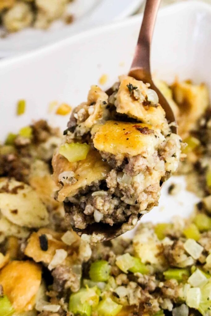 Close-up of a spoonful of baked sage and sausage herb stuffing showing golden bread cubes, sausage, celery, and herbs.