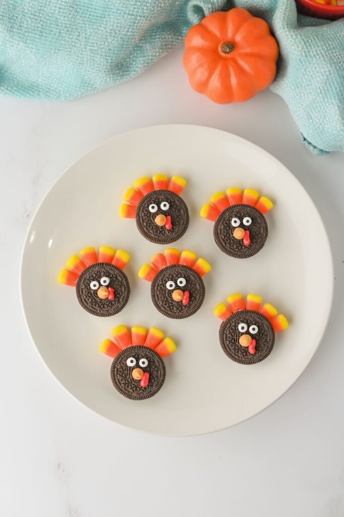 Overhead view of Turkey Oreo Cookies decorated with candy corn feathers, candy eyes, and red frosting wattles on a white plate with a small pumpkin and blue napkin.