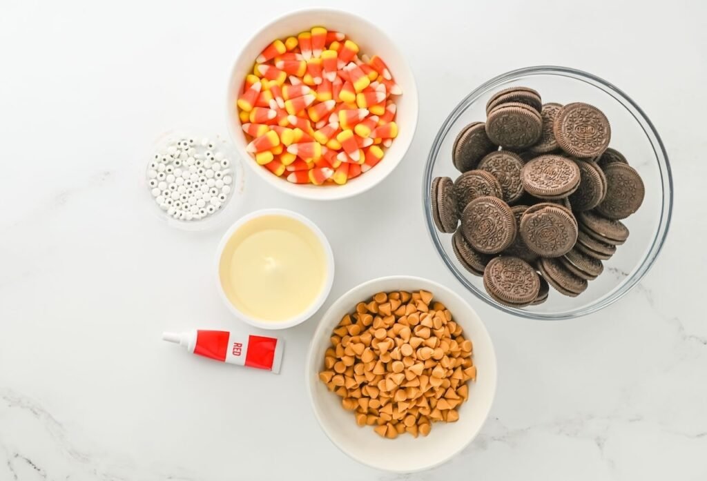 Ingredients for Turkey Oreo Cookies including Oreos, candy corn, frosting, butterscotch chips, and candy eyes arranged in bowls on a white surface.