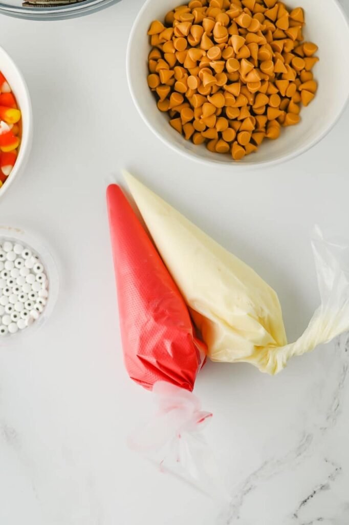 Red and vanilla frosting in piping bags next to bowls of butterscotch chips, candy corn, and candy eyes.