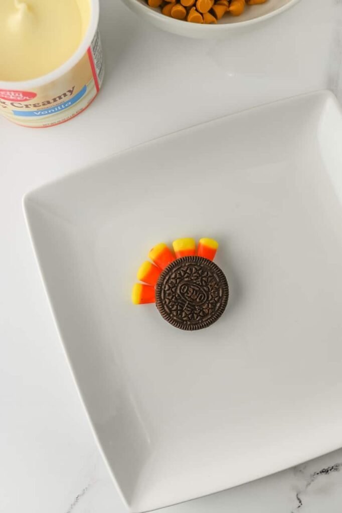 Oreo cookie with candy corn pieces inserted to form colorful tail feathers on a white plate.