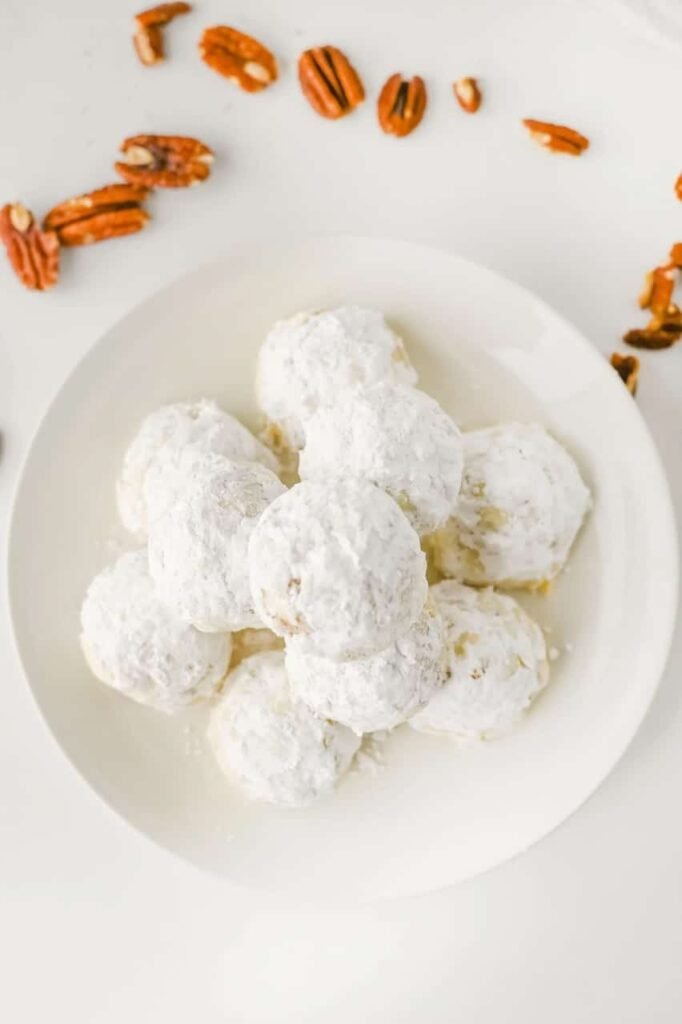 Overhead view of pecan snowball cookies dusted in powdered sugar on a white plate, surrounded by scattered pecans.