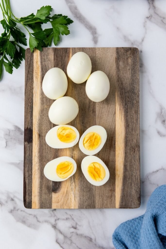 Overhead view of peeled hard-boiled eggs, some halved to show the yolks, arranged on a wooden cutting board with parsley on the side.