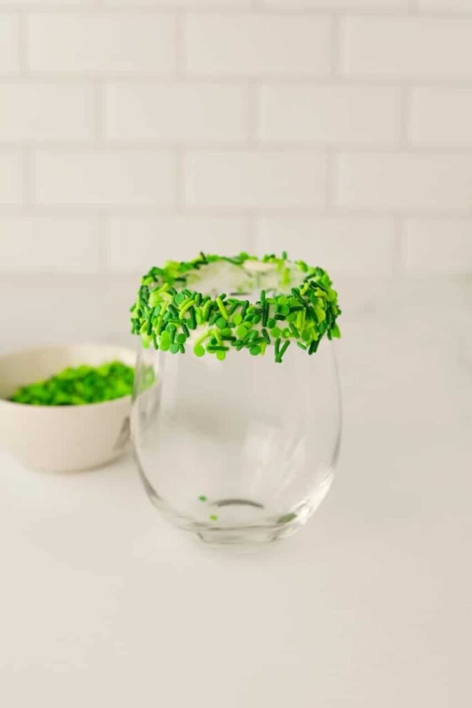 Glass rim coated in frosting and dipped in green sprinkles, prepared for serving a festive Grinch milkshake.