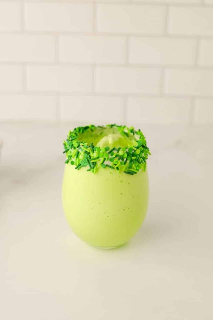 Creamy green holiday shake poured into a glass with a frosting and green sprinkle rim, ready for festive toppings.