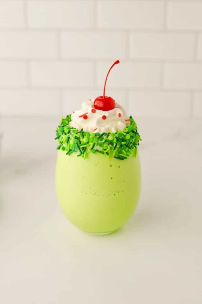 Festive drink topped with whipped cream, red sprinkles, and a cherry, served in a green sprinkle-rimmed glass.