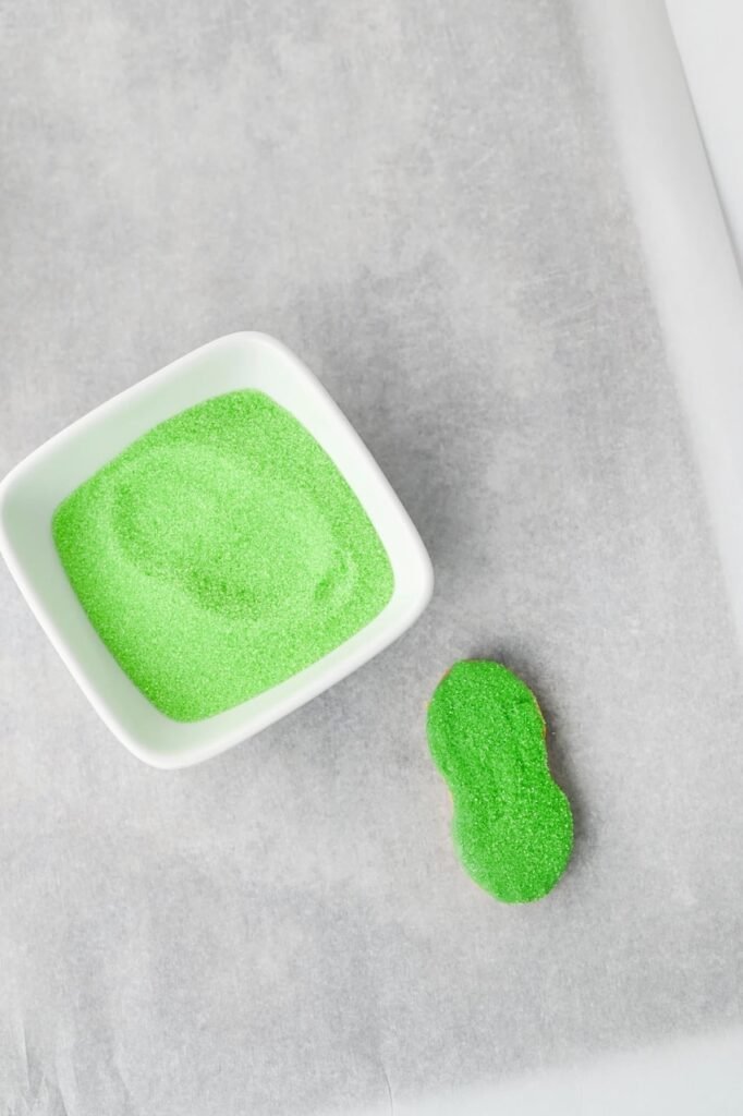 Dipped Nutter Butter cookie coated in green sanding sugar beside a small white bowl of sugar for Grinch Nutter Butter cookies.