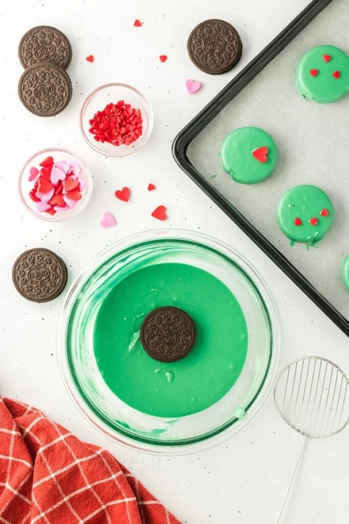 Dipping Oreo cookie into melted green candy coating with red heart sprinkles nearby and finished setting on tray.