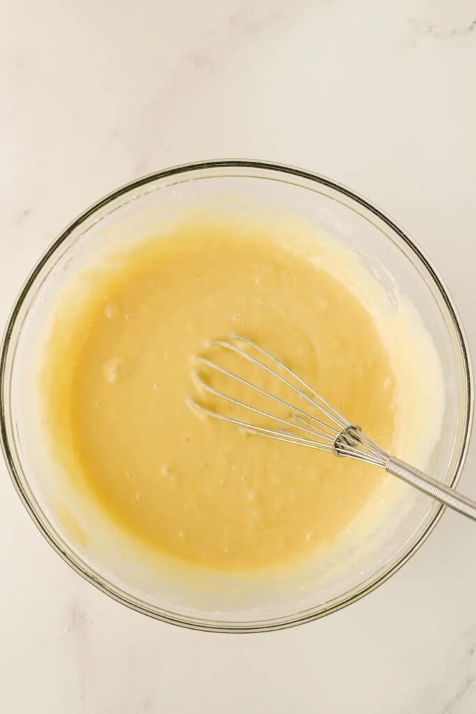 Glass mixing bowl filled with smooth yellow cake batter and a metal whisk on a white marble surface, step one of making a Grinch poke cake.