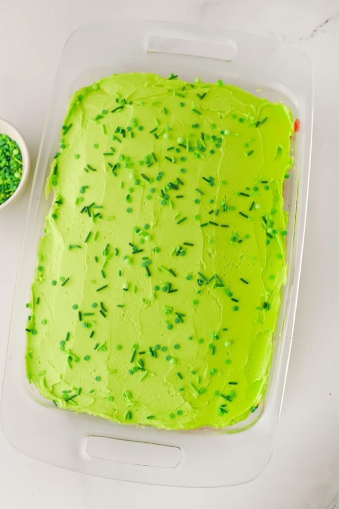 Grinch poke cake topped with smooth green buttercream frosting and festive green sprinkles in a glass baking dish, ready to serve as a kid-friendly Christmas dessert.