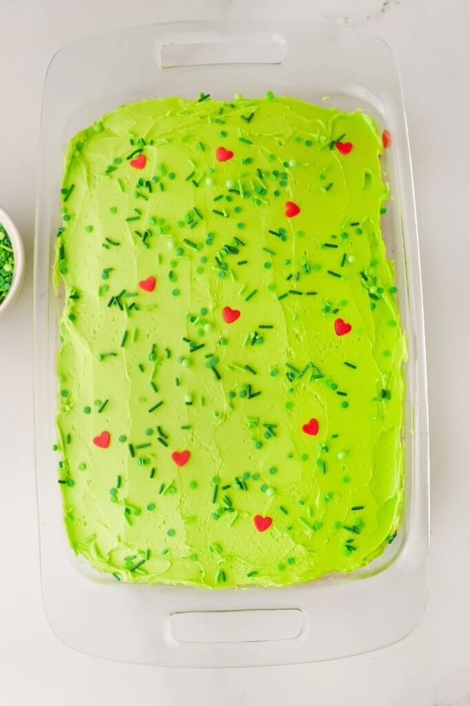 Grinch poke cake topped with green buttercream frosting, festive green sprinkles, and red heart decorations in a glass baking dish, a fun kid-friendly Christmas dessert.