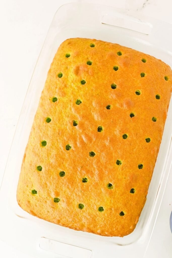 Freshly baked cake in a glass pan with evenly spaced holes poked across the top, ready to be filled with red cheesecake pudding.