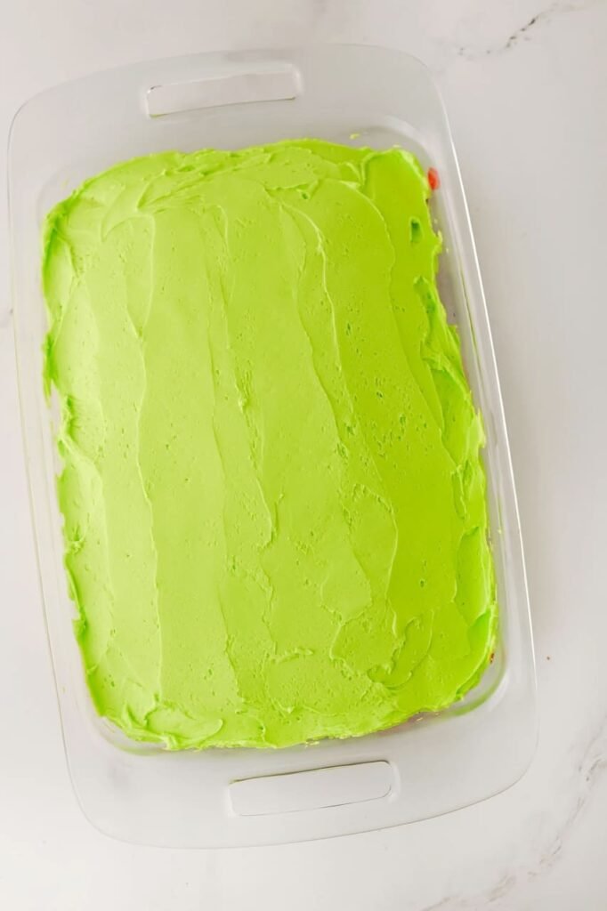 cake covered with smooth, fluffy green buttercream frosting in a glass baking dish, ready for holiday sprinkles and decorations.