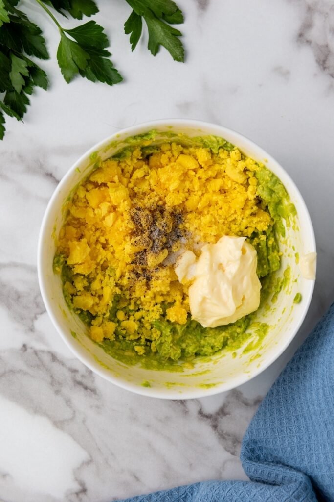 Bowl with mashed avocado, crumbled egg yolks, mayonnaise, salt, and pepper arranged before mixing.