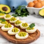 Avocado deviled eggs topped with paprika and parsley on a wooden platter, with fresh avocados, eggs, and herbs in the background.