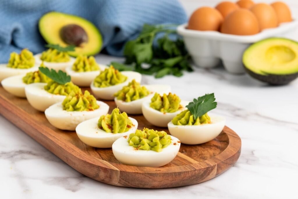 Tray of avocado deviled eggs garnished with paprika and parsley, with fresh avocados, eggs, and herbs in the background.