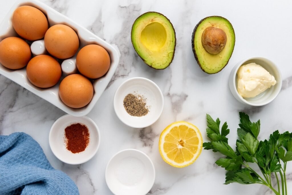 Overhead view of ingredients for avocado deviled eggs, including halved avocados, eggs, lemon, parsley, spices, and seasonings on a marble surface.