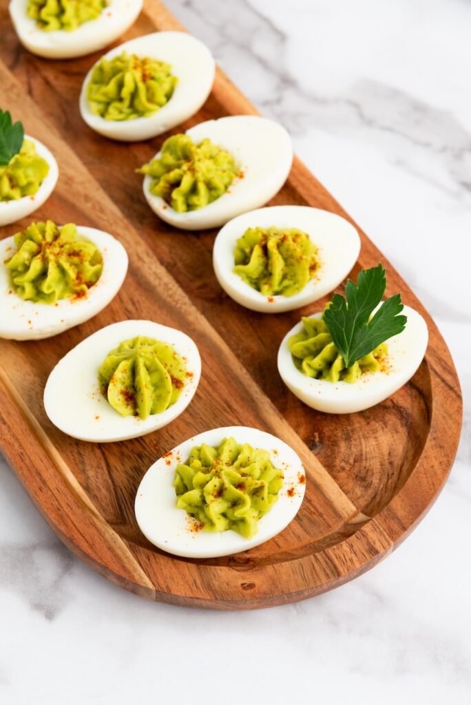 Avocado deviled eggs arranged on a wooden serving platter, topped with paprika and fresh parsley.