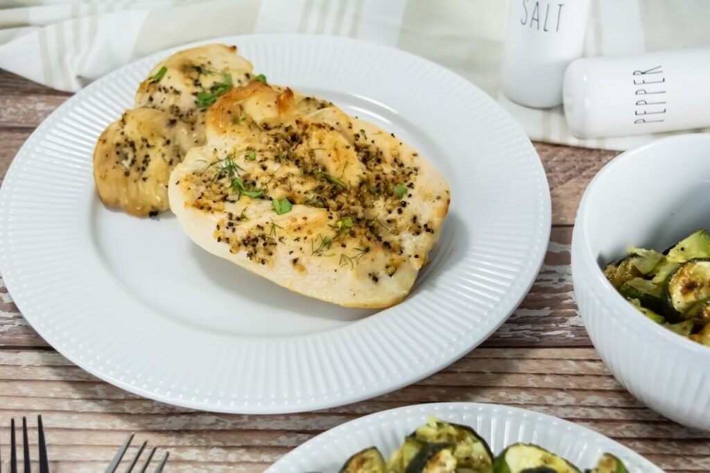 Side angle view of broiled chicken breasts on a white plate with roasted zucchini, seasoned with garlic, black pepper, and herbs.