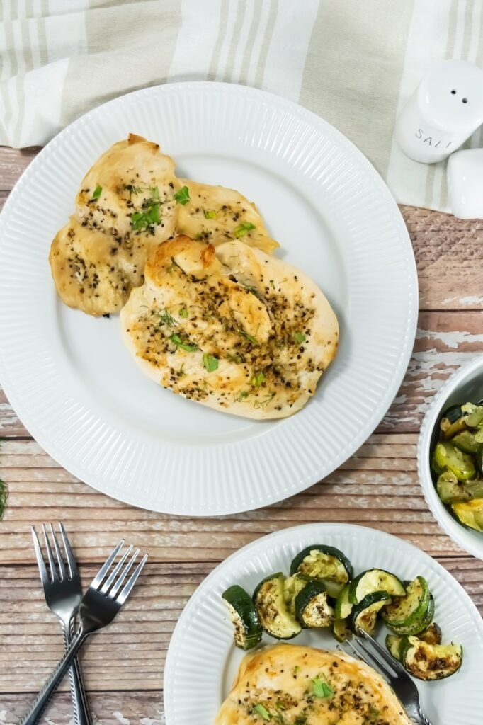 Overhead view of broiled chicken breasts on white plates with roasted zucchini and herbs, styled on a rustic wood table for an easy healthy dinner.