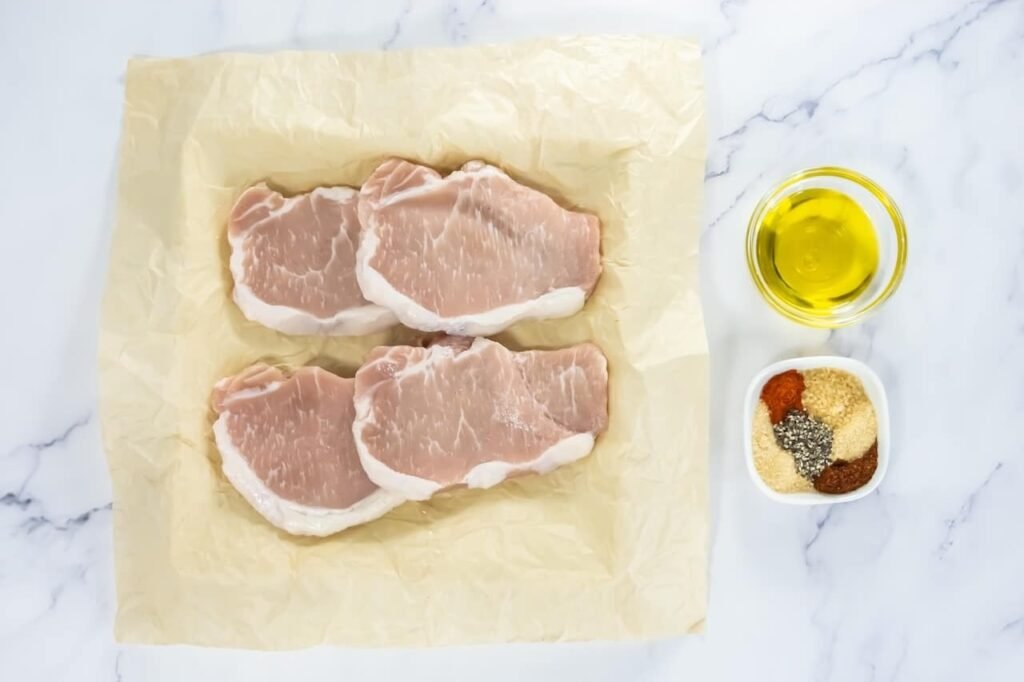 Raw pork chops on parchment paper with a bowl of olive oil and spices ready for broiling.
