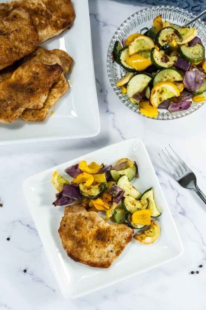 Flat lay of broiled pork chops with roasted zucchini, squash, red onion, and bell pepper served on white plates.