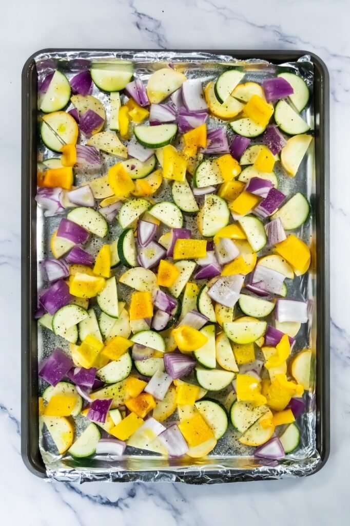 Chopped zucchini, yellow squash, red onion, and yellow bell pepper seasoned with salt, pepper, and olive oil on a foil-lined baking sheet.
