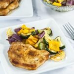 A serving of broiled pork chop with roasted zucchini, squash, onions, and bell peppers on a white plate.