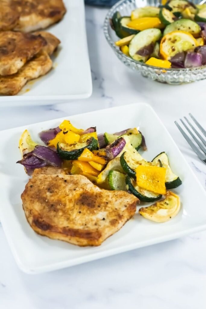 A serving of broiled pork chop with roasted zucchini, squash, onions, and bell peppers on a white plate.