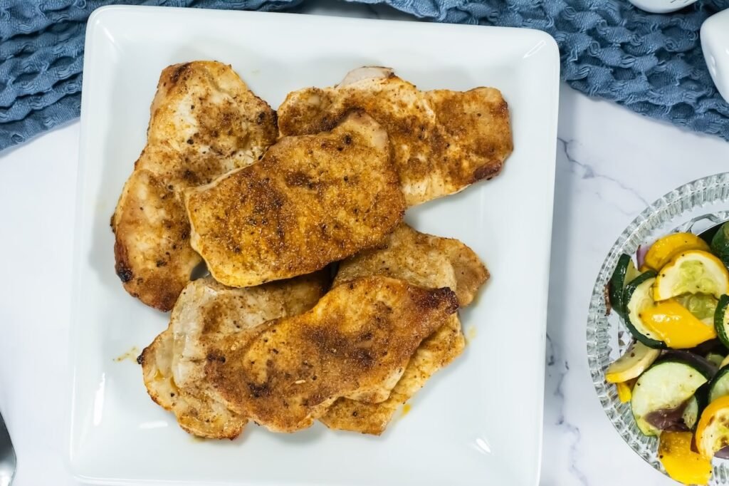 Overhead view of golden-brown broiled pork chops served on a white plate with a side of roasted zucchini, squash, and onions.