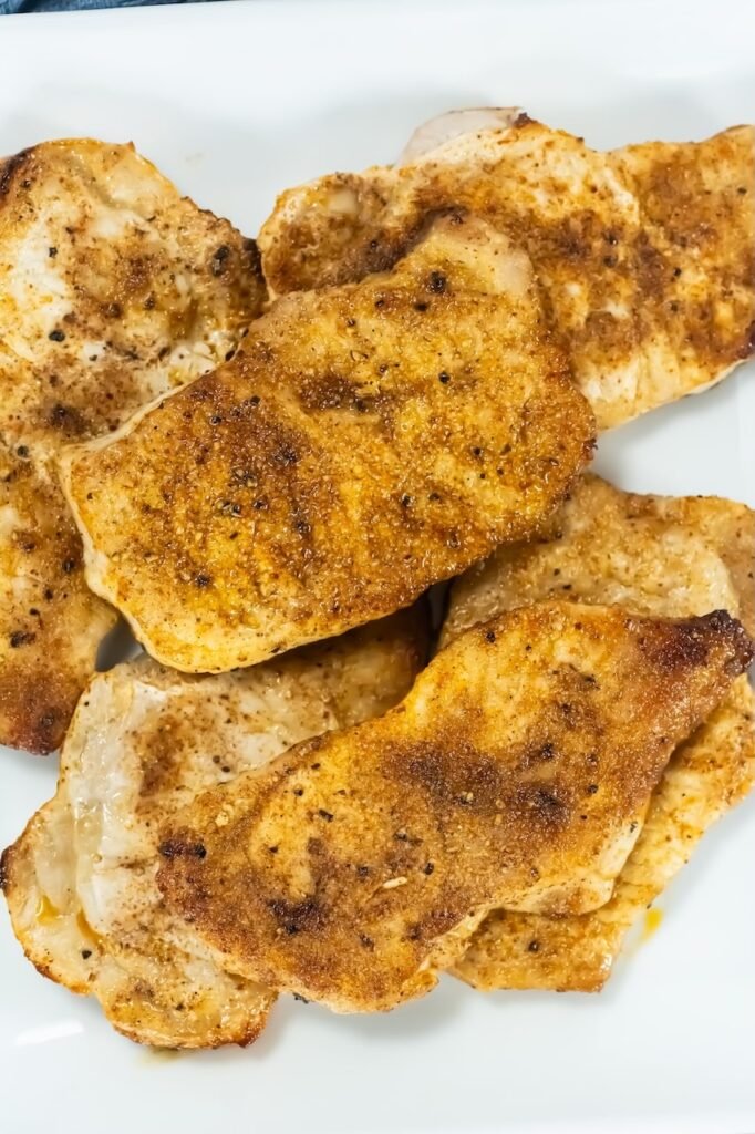 Close-up of golden-brown broiled pork chops on a white plate, showing crispy, seasoned edges.