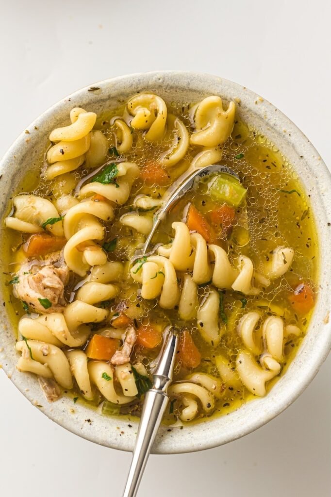 Top-down view of a bowl of chicken noodle soup with spiral noodles, carrots, celery, and chicken in a golden broth, with a spoon resting inside.