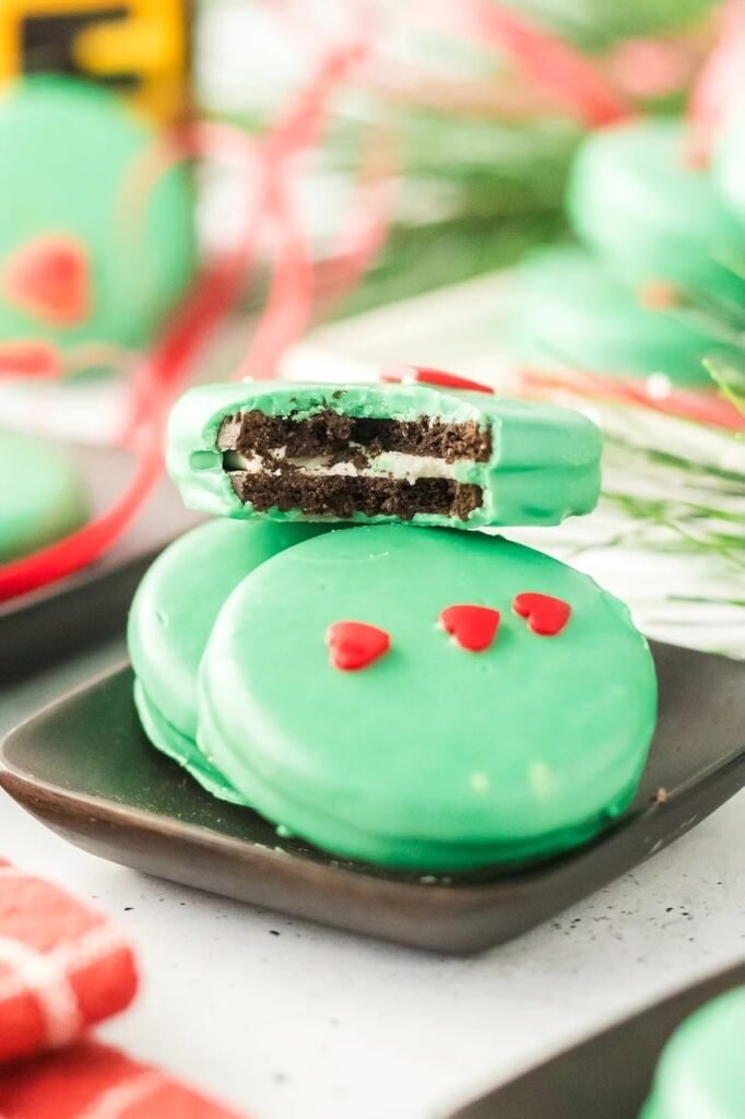 Close-up of Grinch Oreo cookie with a bite taken out, showing Oreo filling and green candy coating topped with red heart sprinkles.