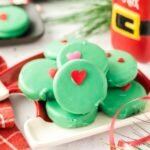 Stack of green Grinch Oreos with red and pink heart sprinkles on a festive tray beside Santa’s milk bottle.