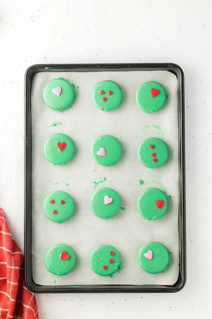 Green candy-coated topped with red and pink heart sprinkles setting on a parchment-lined baking tray.