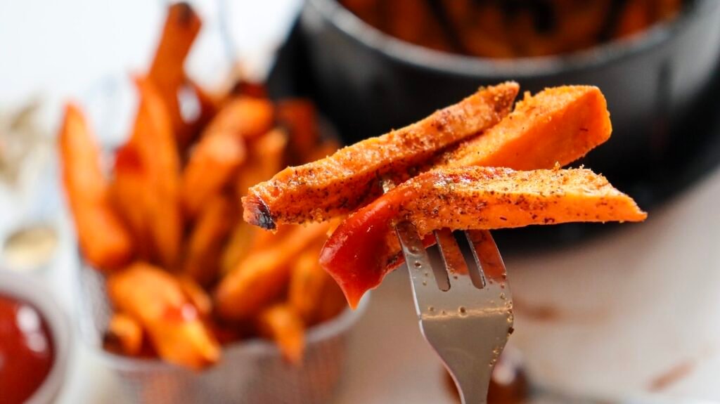 Close-up view of crispy air fryer sweet potato fries resting on a fork, lightly browned with seasoned edges, dipped in ketchup, with more fries blurred in the background.
