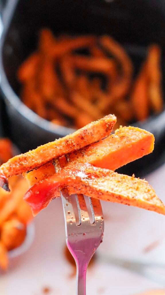 Close-up view of crispy air fryer sweet potato fries resting on a fork, lightly browned with visible seasoning and crisp edges, with the air fryer basket and more fries blurred in the background.