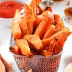Close-up of crispy air fryer sweet potato fries stacked in a small metal basket, lightly browned with seasoned edges, ready to serve with dipping sauce in the background.