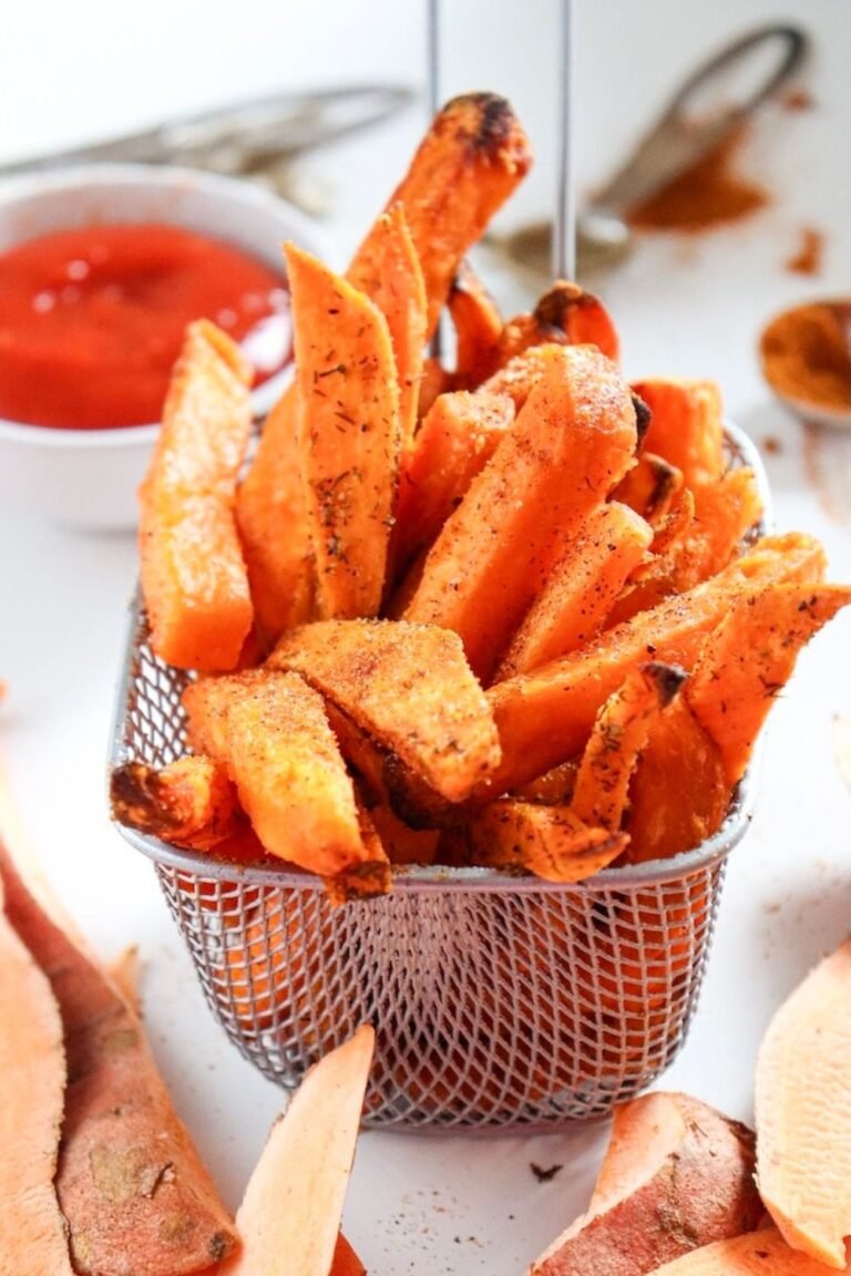 Close-up of crispy air fryer sweet potato fries stacked in a small metal basket, lightly browned with seasoned edges, ready to serve with dipping sauce in the background.