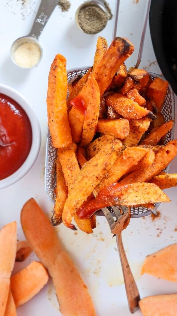 Overhead close-up of crispy air fryer sweet potato fries being lifted from a small wire basket, with seasoned edges, dipping sauce nearby, and spices scattered on the surface.