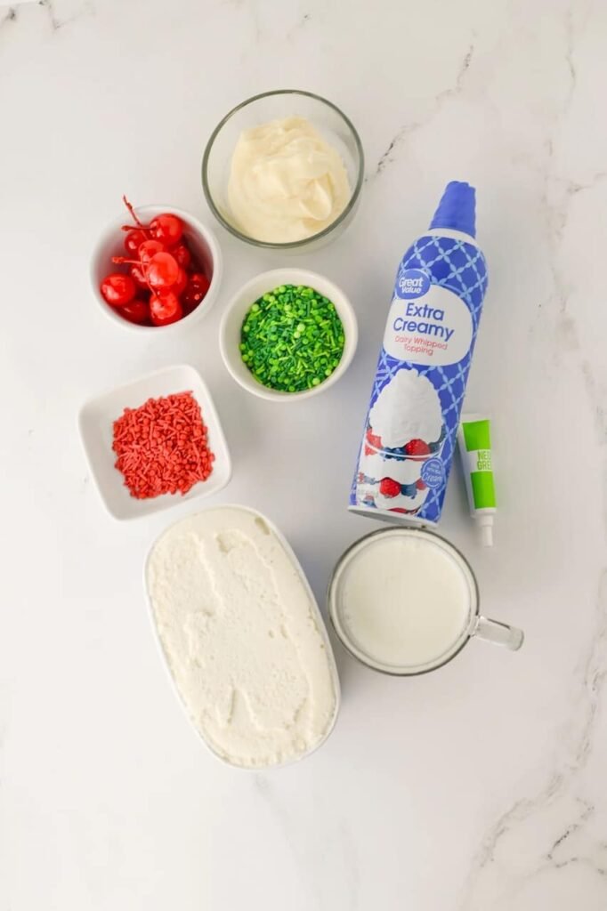 Overhead view of ingredients for a Grinch milkshake, including vanilla ice cream, milk, whipped cream, green food coloring, red and green sprinkles, and cherries on a marble countertop.