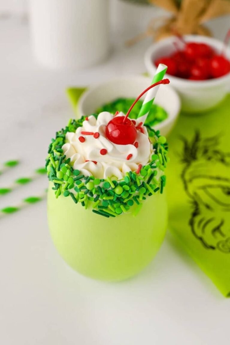 Close-up of a festive Grinch milkshake topped with whipped cream, red sprinkles, and a cherry, served with a green striped straw and holiday napkin.