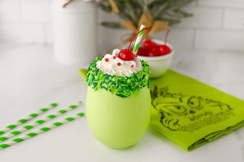 Festive Grinch milkshake topped with whipped cream, red sprinkles, and a cherry, served in a green sprinkle-rimmed glass with a striped straw on a holiday napkin.