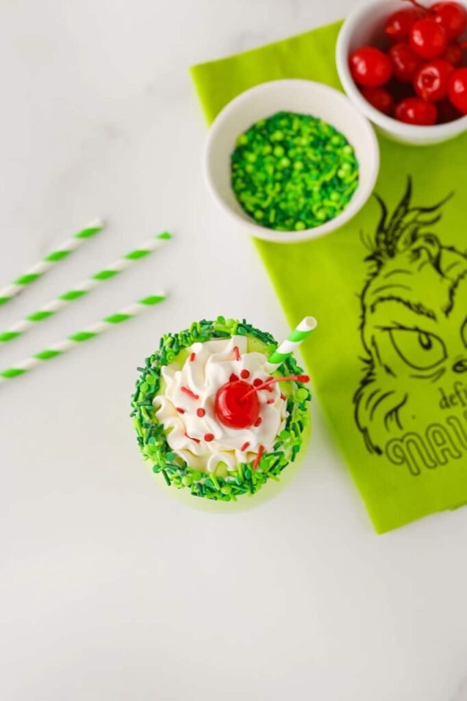 Overhead view of a festive Grinch milkshake topped with whipped cream, red sprinkles, and a cherry, surrounded by striped straws, sprinkles, and holiday decor.