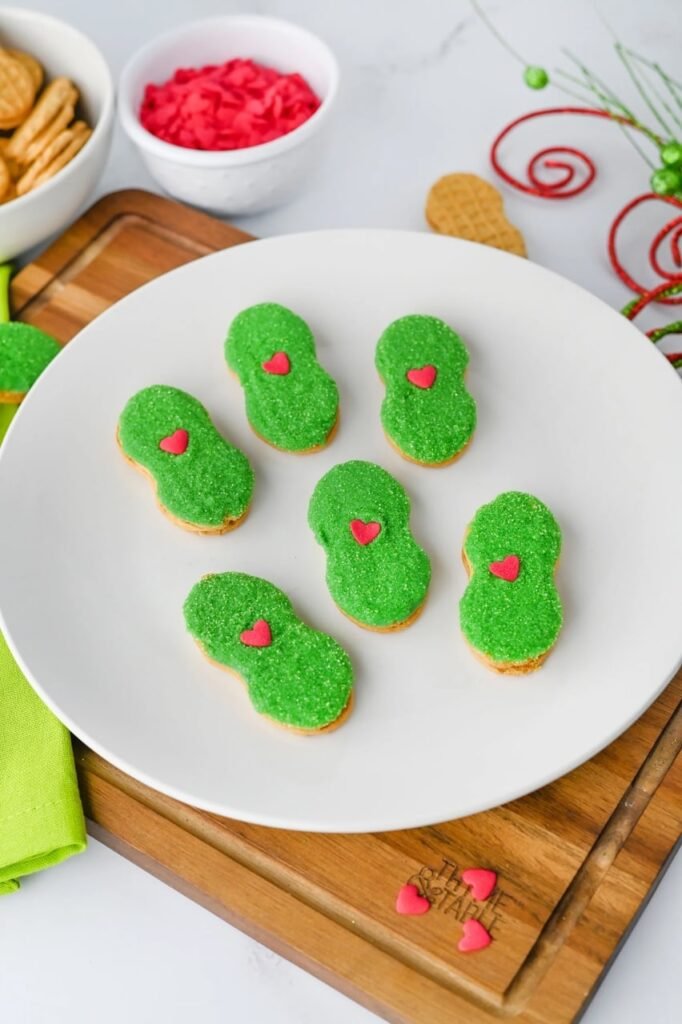 Plate of Grinch Nutter Butter cookies with green sanding sugar and red hearts on a wooden board — an easy no-bake Christmas cookie idea.